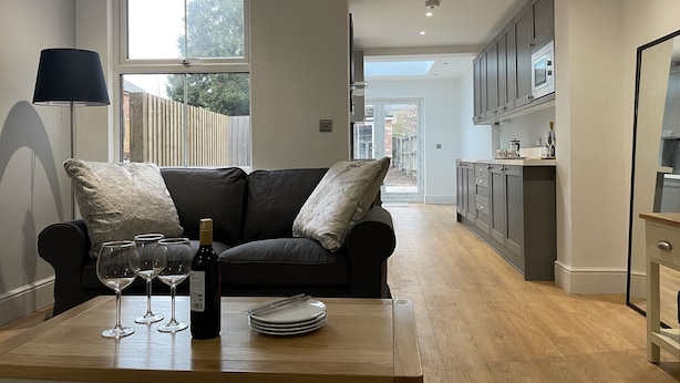 A modern kitchen with dark cabinets, a wooden countertop, and a dining area with a black sofa and a table set with wine glasses and plates. - Beautiful Townhouse in Stratford Upon Avon (Stratford-upon-Avon)