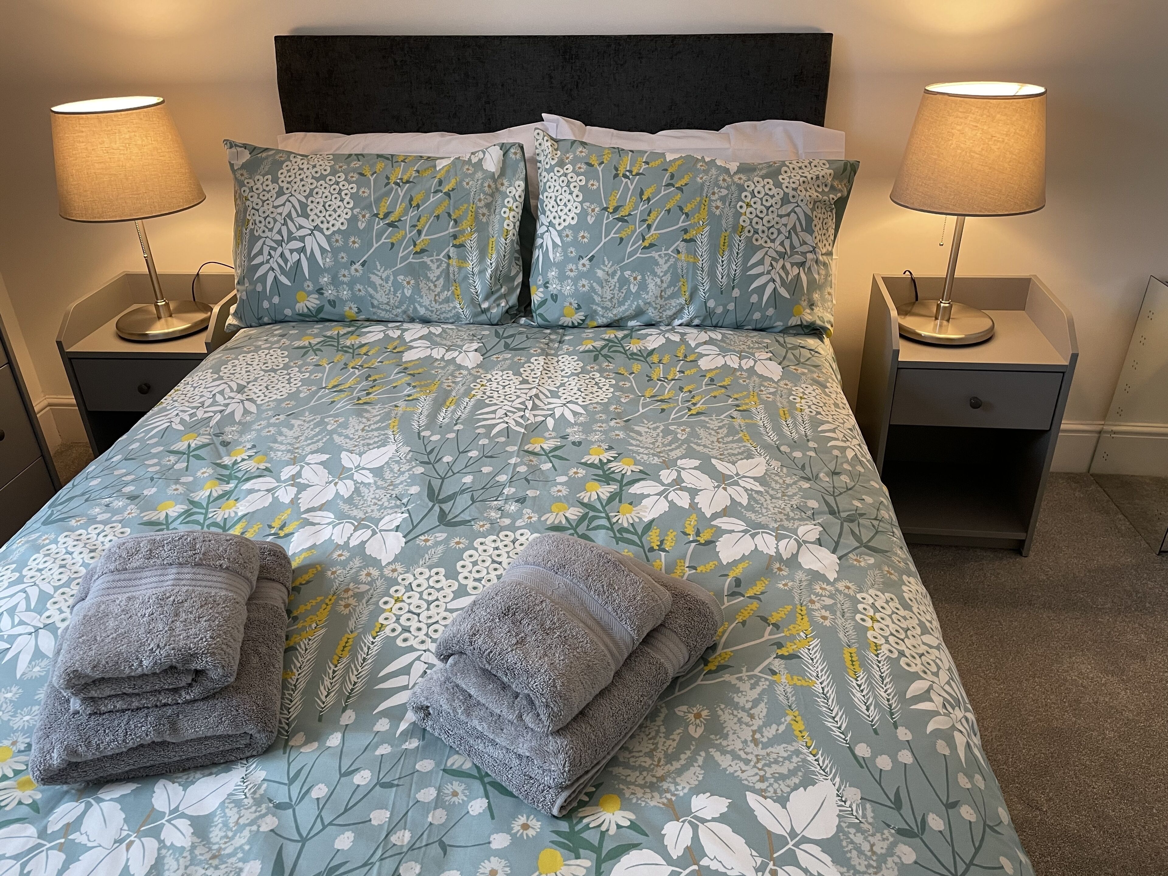 Ein ordentlich bezogenes Bett mit floralem Bettzeug, zwei Nachttischlampen und zwei gefaltete Handtücher auf dem Bett.