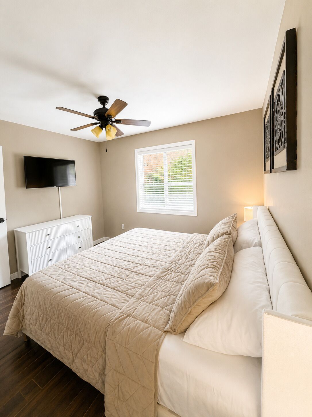 Een slaapkamer met een bed, een plafondventilator, een televisie, een raam en een witte dressoir.