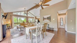 A spacious living room with a high ceiling, wooden beams, a dining area with a white table and chairs, and large windows offering a view of the outdoors. - Treetop & Waterfront! Quiet Cove, Hot Tub, Kayaks, Game Room and more (Moneta)
