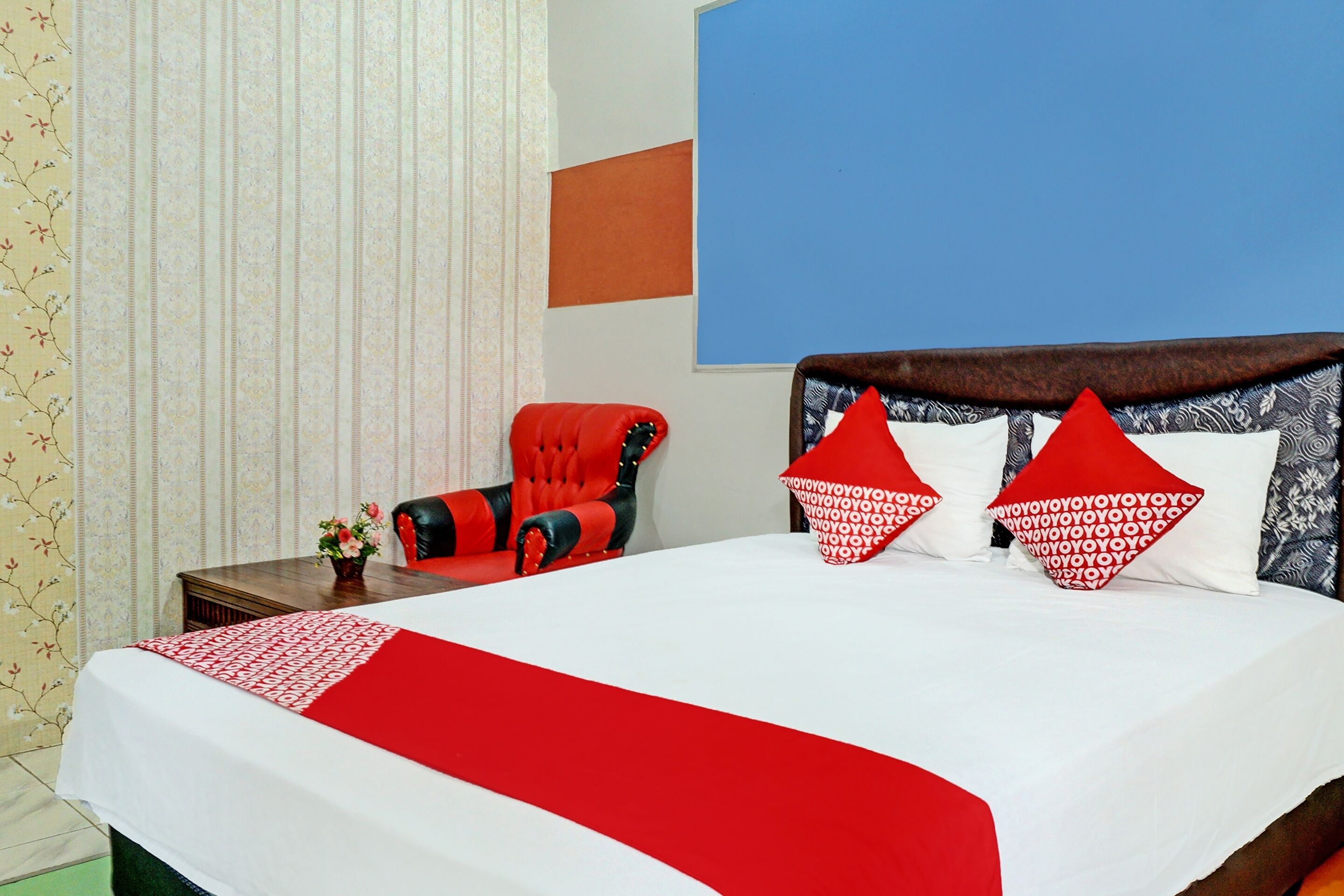 A hotel room with a large bed, red and white bedding, a red armchair, and a blue accent wall.