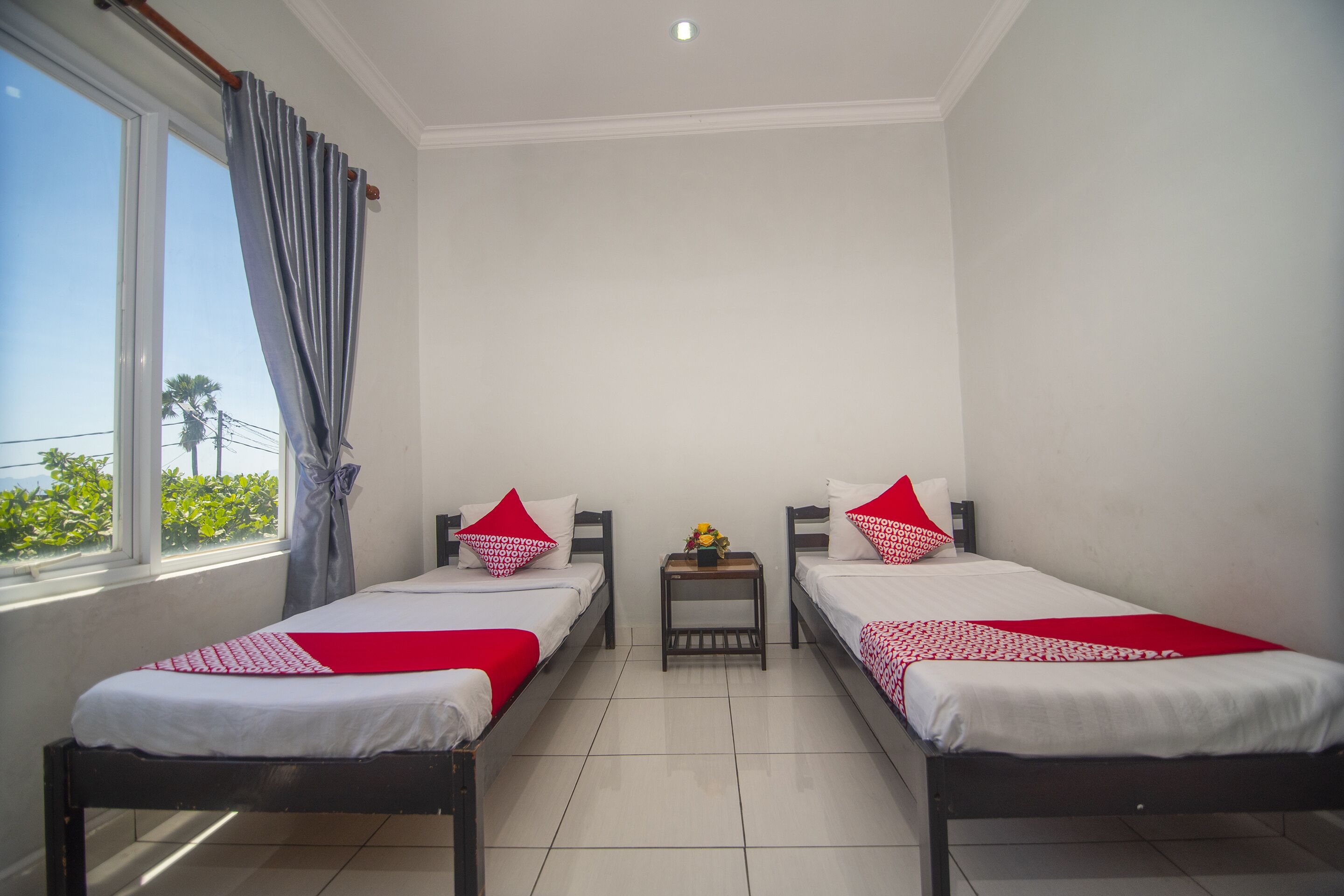 A room with two single beds, a small table with a vase, and a window with curtains.