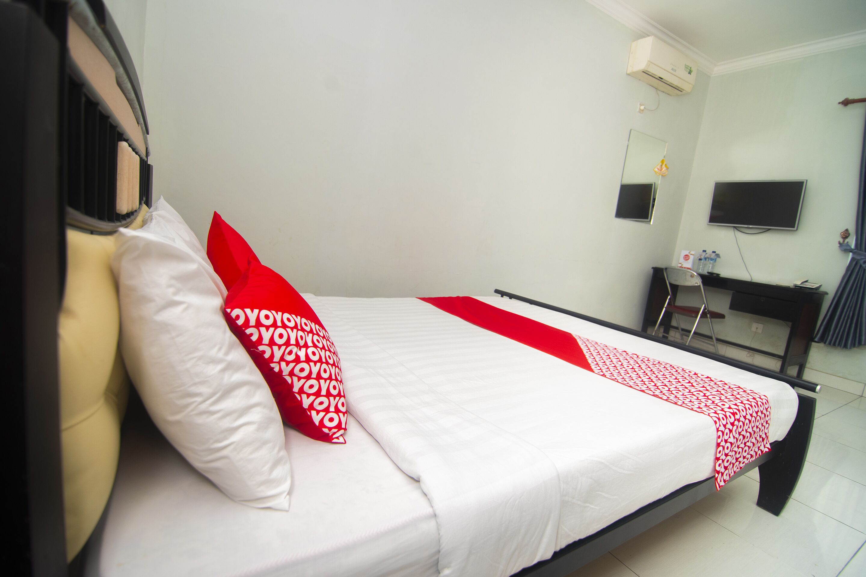 A neatly made bed with white and red bedding, a desk with a chair, a flat-screen TV, and an air conditioning unit on the wall.