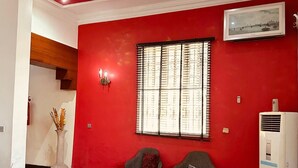 A living room with red walls, two grey sofas, a red armchair, and a coffee table. There is a window with blinds, a painting on the wall, and a white air conditioning unit. - BATES GARDEN GUEST HOUSE (Lekki)