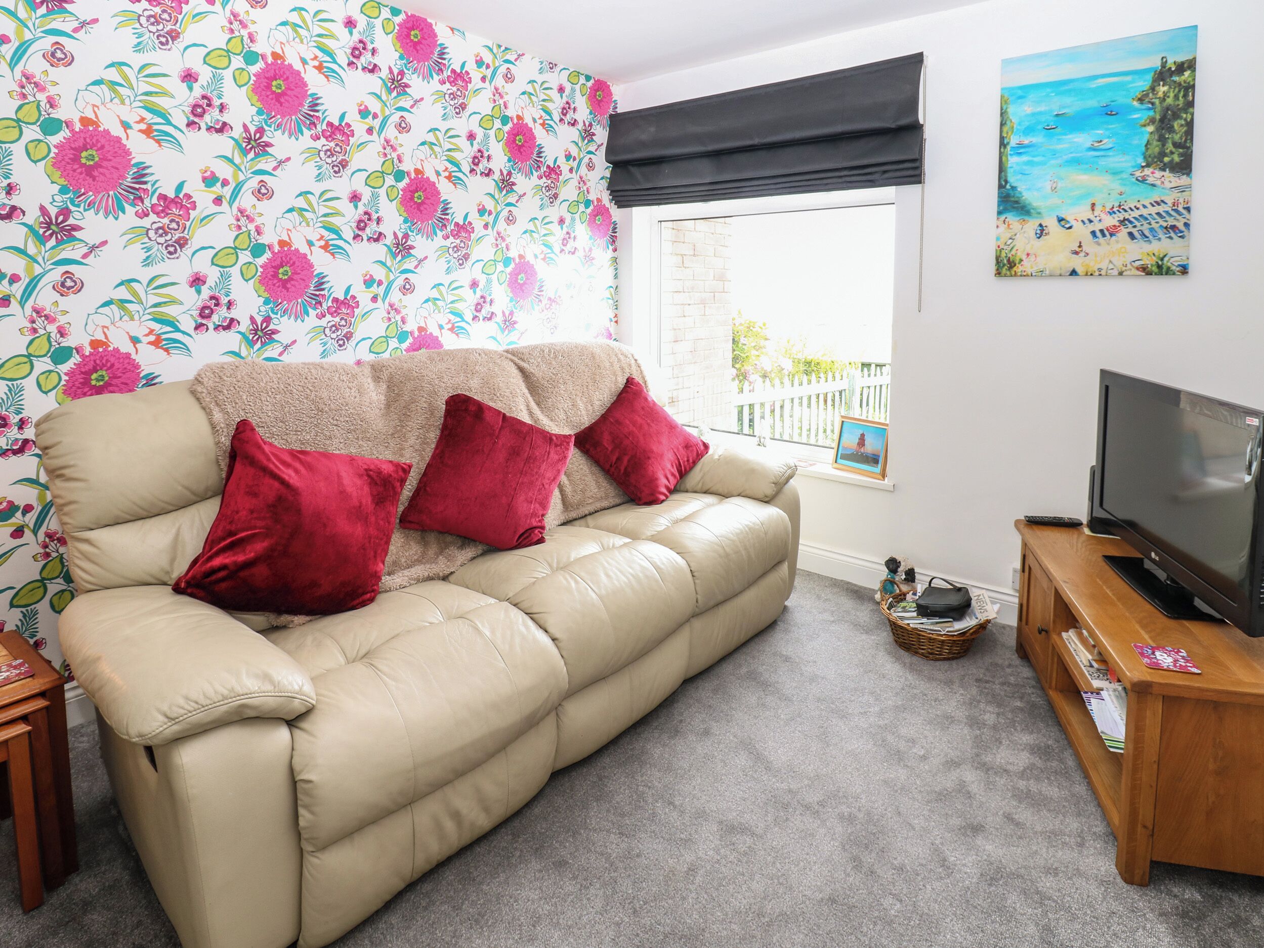 A living room with a beige sofa, red cushions, a TV on a wooden stand, a window with a view of a beach, and a floral wallpaper.