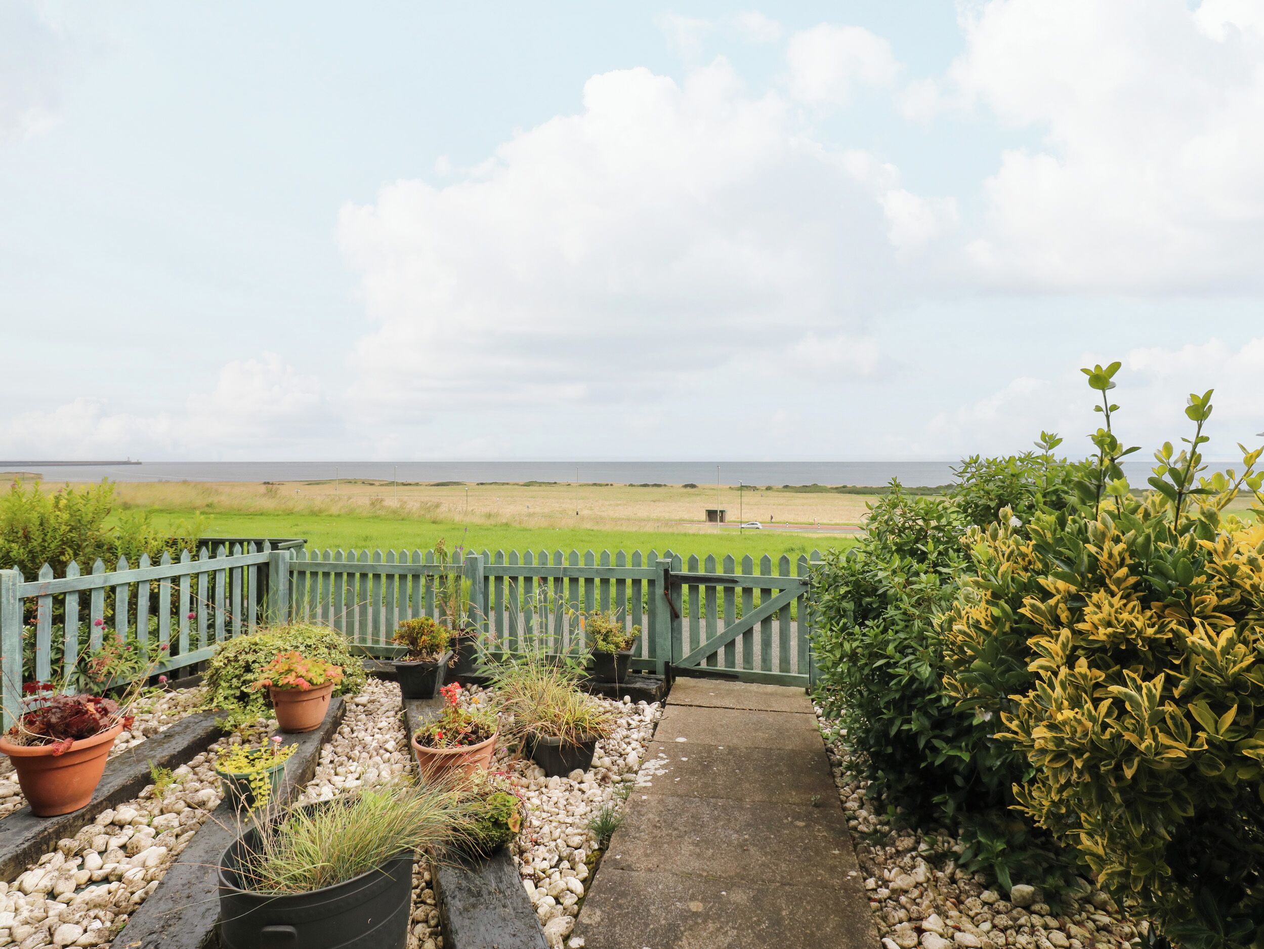 A pathway leading to a green fence overlooking a grassy field and the sea.