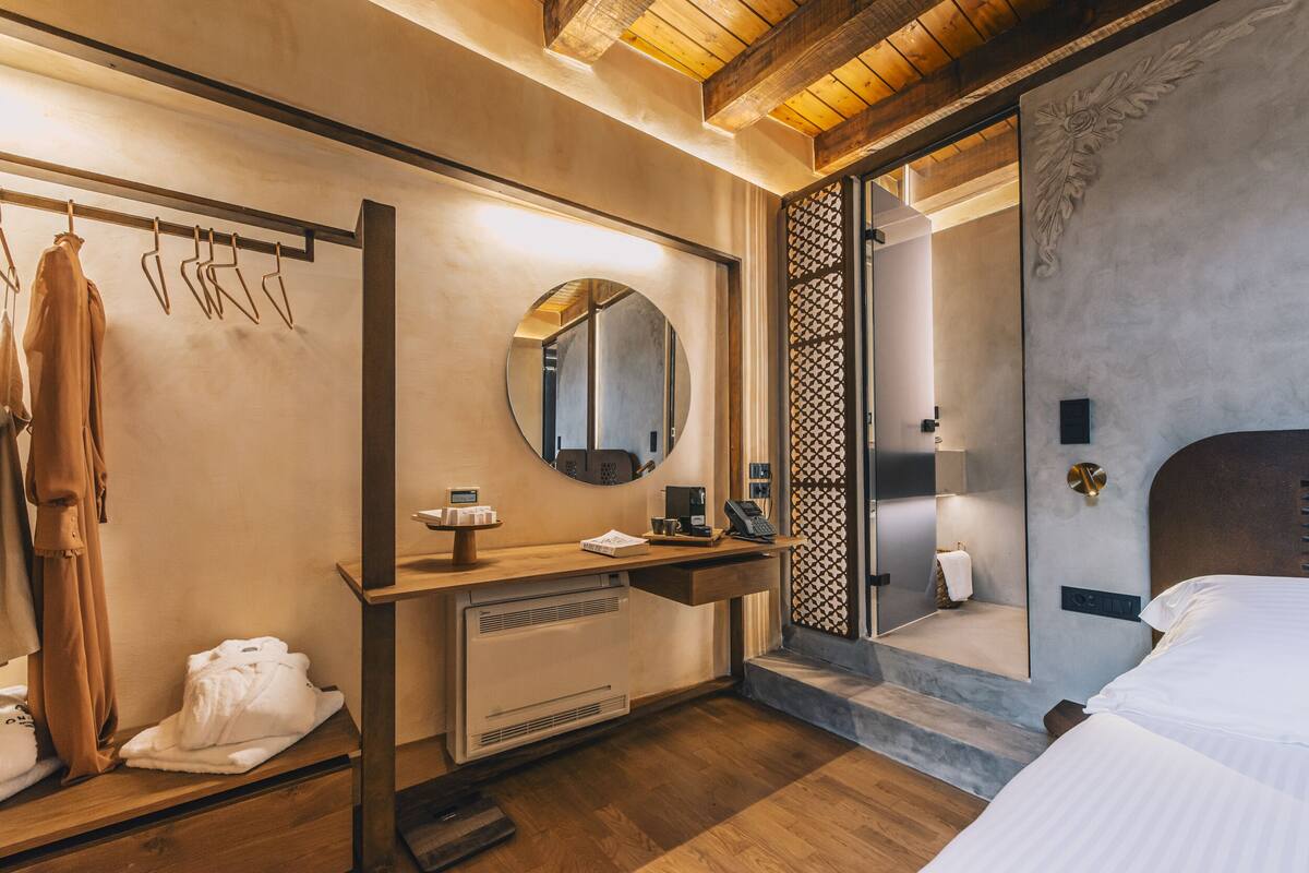 A hotel room with a wooden floor, a round mirror, a bedside table, a bed, a radiator, and a hanging clothes rack.