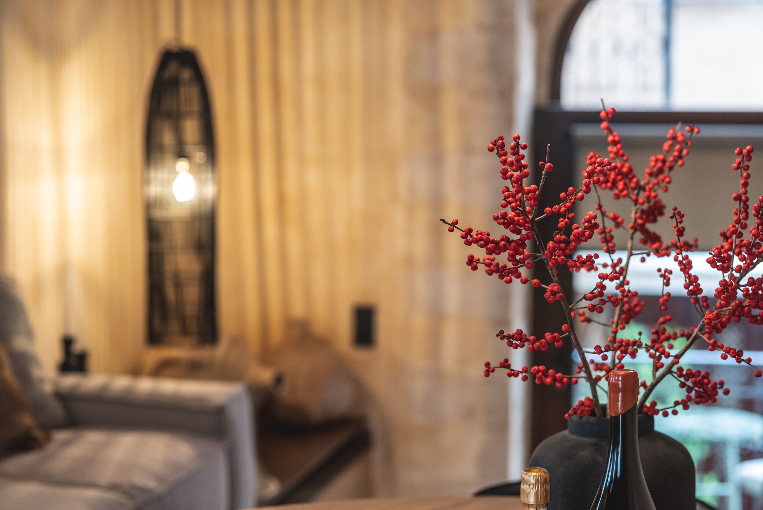 Una habitación con una cama, un florero con bayas rojas y un ventanal con cortinas.