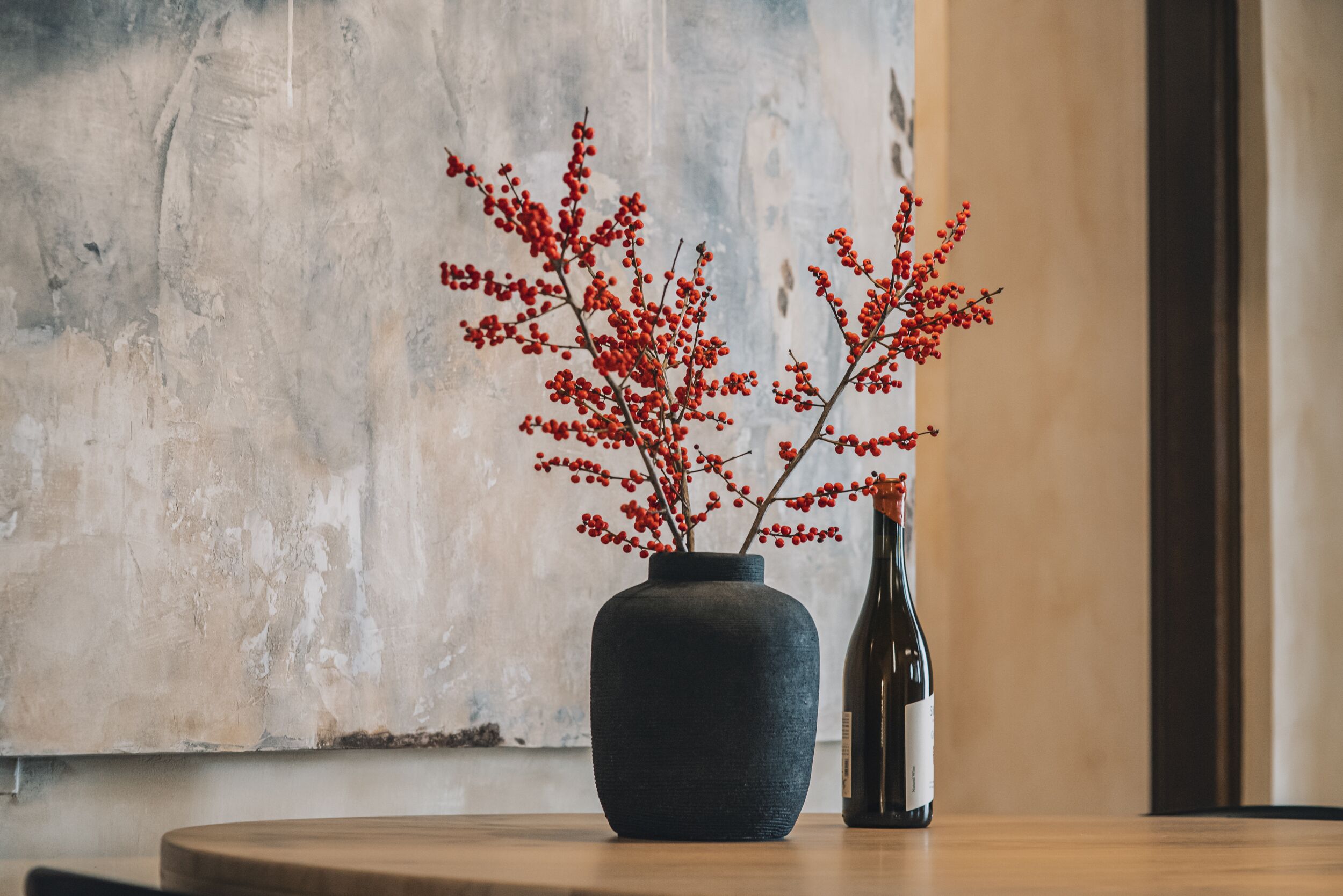 Un vase foncé orné de baies rouges, posé sur une table en bois devant un mur à texture.