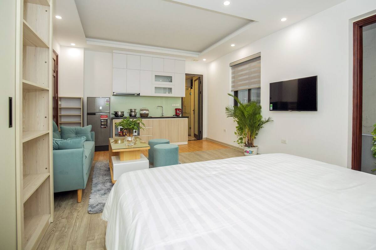 A modern bedroom with a large bed, a kitchenette, a seating area with a sofa and ottoman, and a TV mounted on the wall.