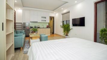Un dormitorio moderno con una cama grande, una kitchenette, una zona de estar con un sofá y reposapiés, y un televisor montado en la pared.
