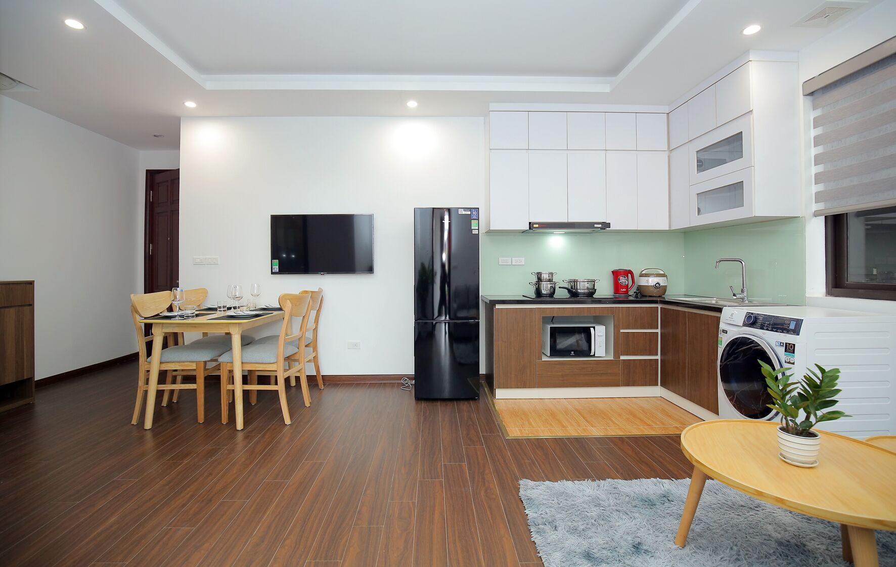 A modern kitchen with a dining area, a black refrigerator, a microwave, a washing machine, and a flat-screen TV.