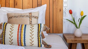 Un letto rifatto con cura, con una testata in legno, cuscini bianchi e un cuscino decorativo colorato. Accanto al letto, un comodino in legno con un vaso di tulipani.