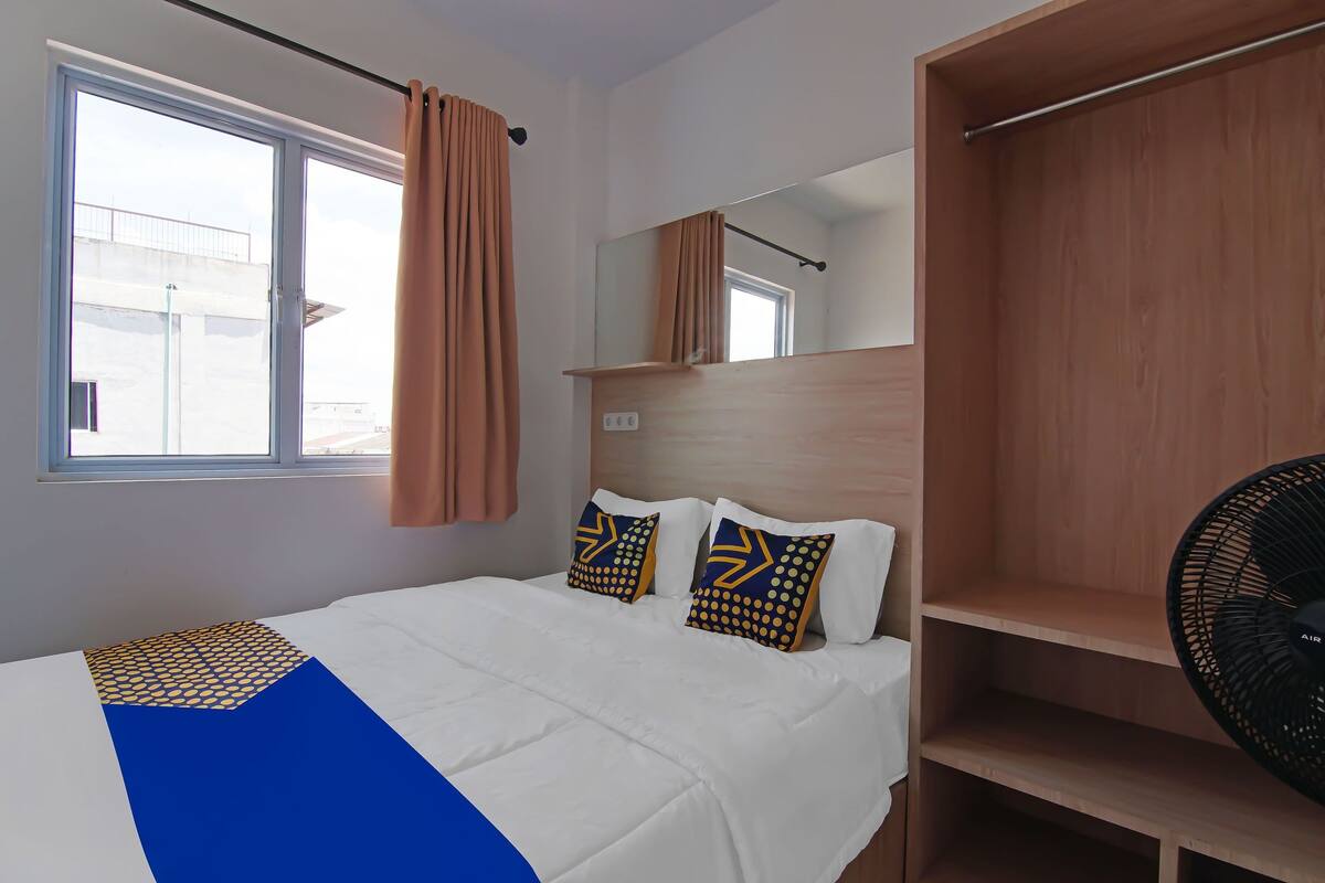 A hotel room with a large window, a bed with patterned pillows, a wooden wardrobe, and a standing fan.