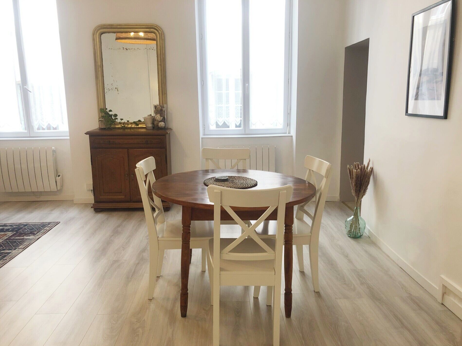 Uma área de refeições com uma mesa de madeira redonda, cadeiras brancas, um espelho e um radiador.