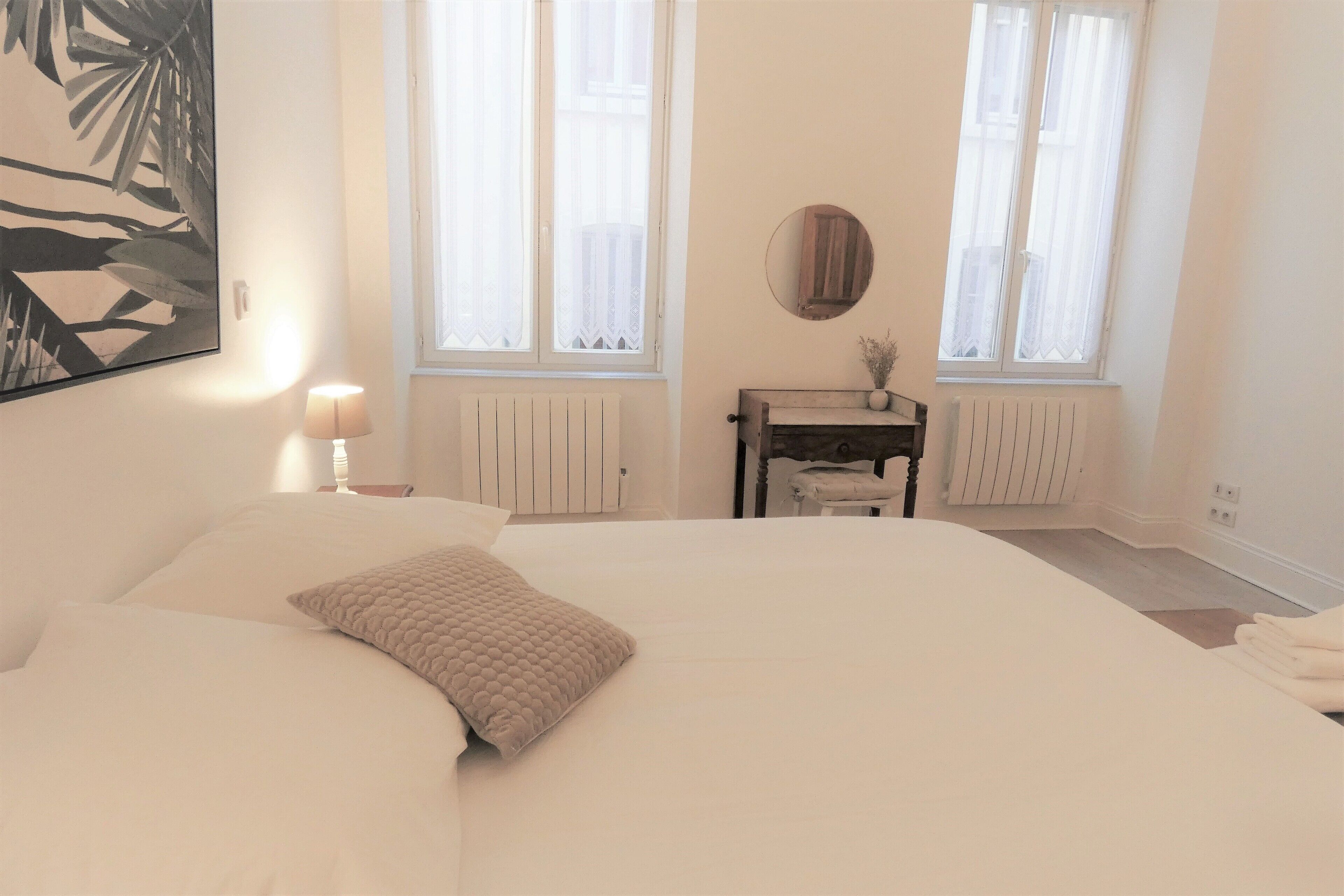 A bedroom with a large bed, a bedside table with a lamp, a mirror, and two windows with sheer curtains.