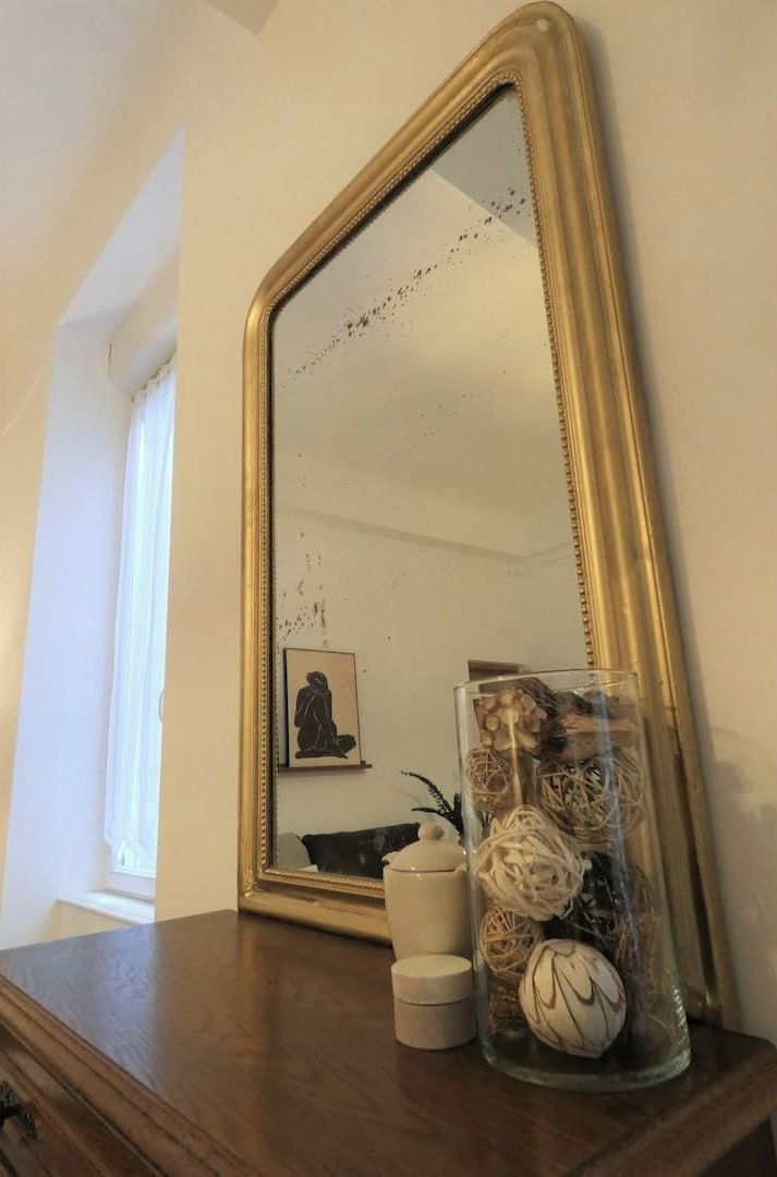 A mirror with a gold frame on a wooden dresser, a small picture frame, and a glass jar with decorative items.