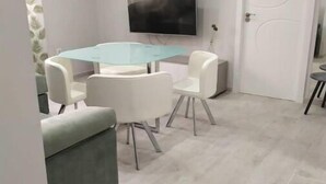 Ein modernes Wohnzimmer mit Esstisch aus Glas, weißen Stühlen, einem grauen Sofa, einem flachbildfernseher an der Wand und einer weißen Tür.