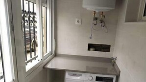 Ein kleiner Waschraum mit Waschmaschine, einer kleinen Ablagefläche und einem Fenster mit Gitterstäben.