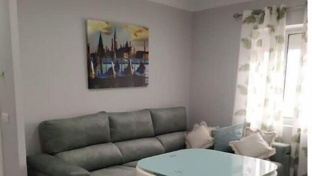 Ein Wohnzimmer mit grauem Sofa, einem Couchtisch mit Glasplatte und weißen Stühlen. An der Wand hängt ein Gemälde und ein Fenster mit durchsichtigen Vorhängen.