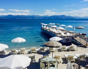 A beachside resort with sun loungers and umbrellas, a pier extending into the sea, and mountains in the background. - New T2, Cap d'Antibes, very comfortable, terrace, air conditioning, parking (Antibes)