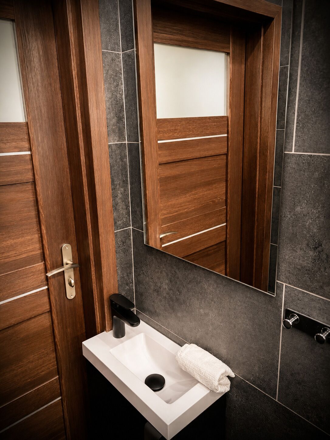 A modern bathroom with a wooden door, a white sink, a mirror, and dark tiled walls.