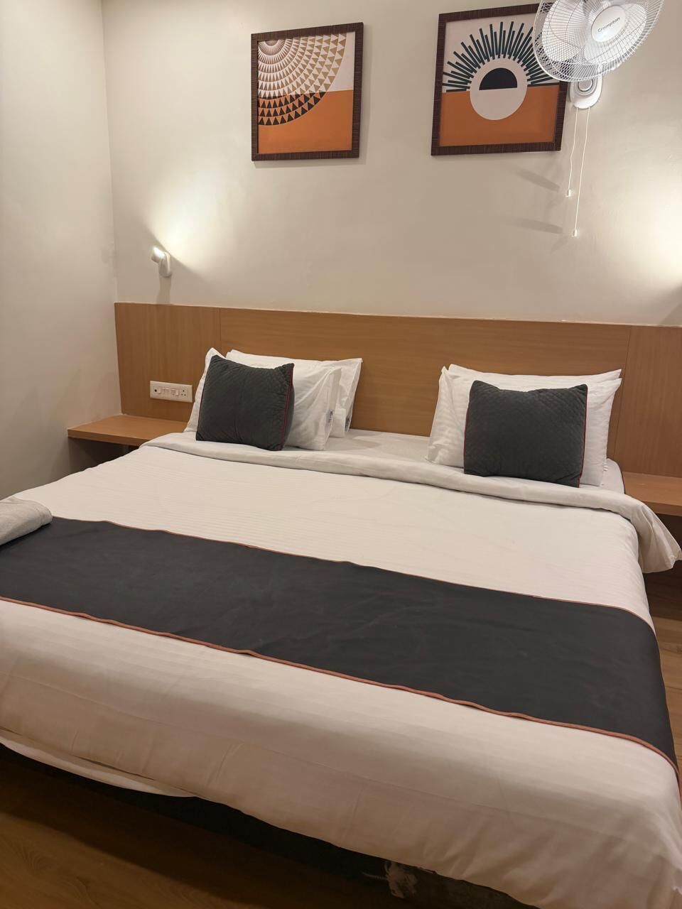 Une chambre d’hôtel comprenant un lit, deux tables de chevet, un ventilateur fixé au mur et deux œuvres d’art encadrées.