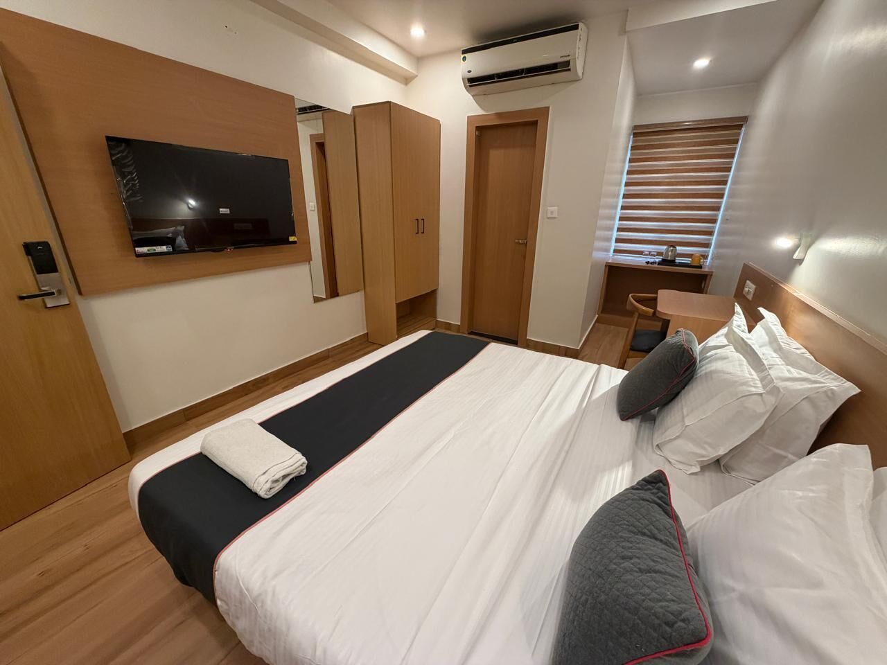A hotel room with a bed, a TV mounted on the wall, a wardrobe, and an air conditioning unit.