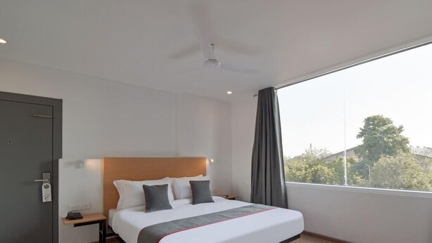 Una habitación de hotel moderna con una cama grande, una mesita de noche, un ventilador de techo y un ventanal con vistas a árboles y edificios.