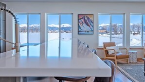 A modern dining area with a large table, chairs, and a view of snow-covered mountains through large windows.