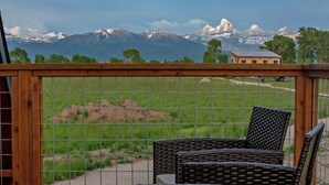 A balcony with wicker chairs, a wooden table, and a view of mountains and a house.