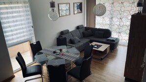 A modern living room with a sectional sofa, a glass dining table, black chairs, and a coffee table. There are framed artworks on the walls, a large window with blinds, and a patterned curtain. - Kumitzblick: A Modern, High-End Vacation Rental (Pichl-Kainisch)