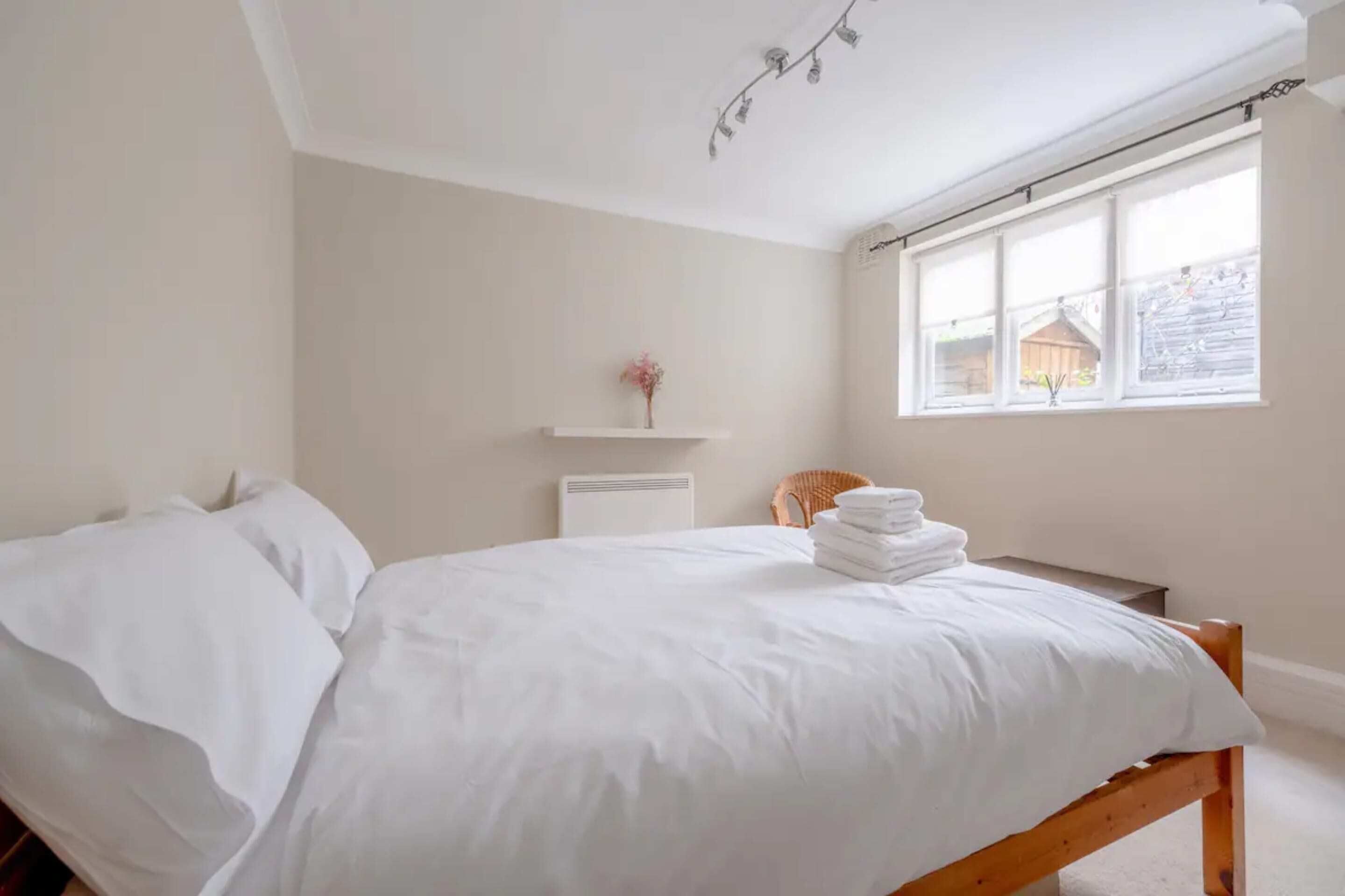 A neatly made bed with white linens in a room with a large window, a small shelf with a vase, and a wicker chair.