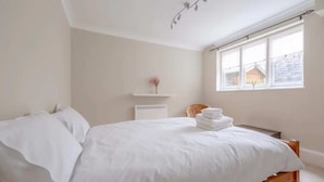 A neatly made bed with white linens in a room with a large window, a small shelf with a vase, and a wicker chair. - Lovely 1 Bedroom With Patio - 10 Mins From Hyde Park (London)