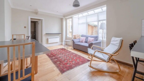 A spacious living area with a dining table, a sofa, a rocking chair, and a large window providing natural light. - Lovely 1 Bedroom With Patio - 10 Mins From Hyde Park (London)