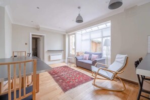 A spacious living area with a dining table, a sofa, a rocking chair, and a large window providing natural light.