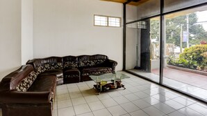 A living room with a brown leather sofa, a glass coffee table, and a large window offering a view of the outdoors. - Hotel O Penginapan Aina Syariah (Lebaho Ulaq)