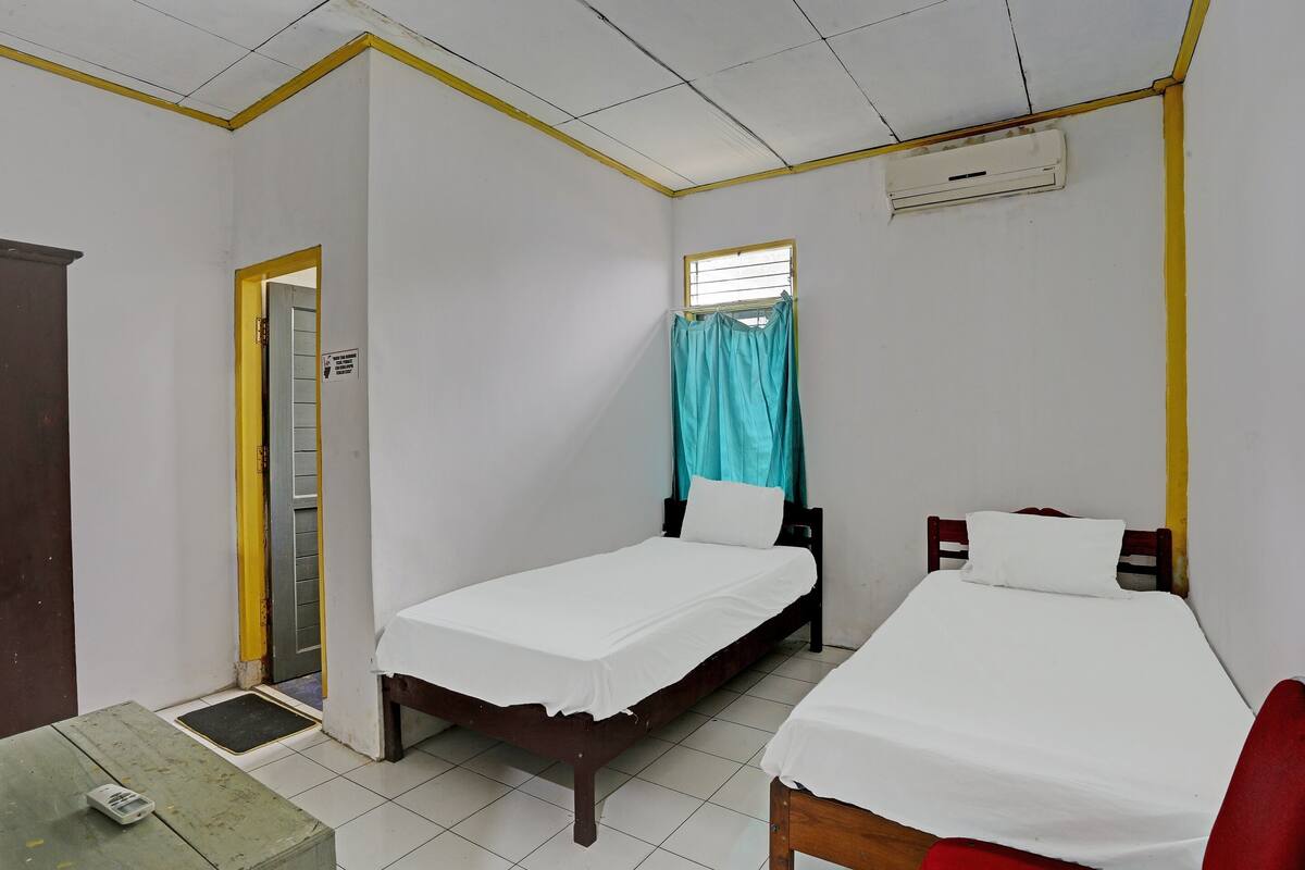A room with two single beds, a small table, a chair, a window with curtains, and an air conditioning unit.