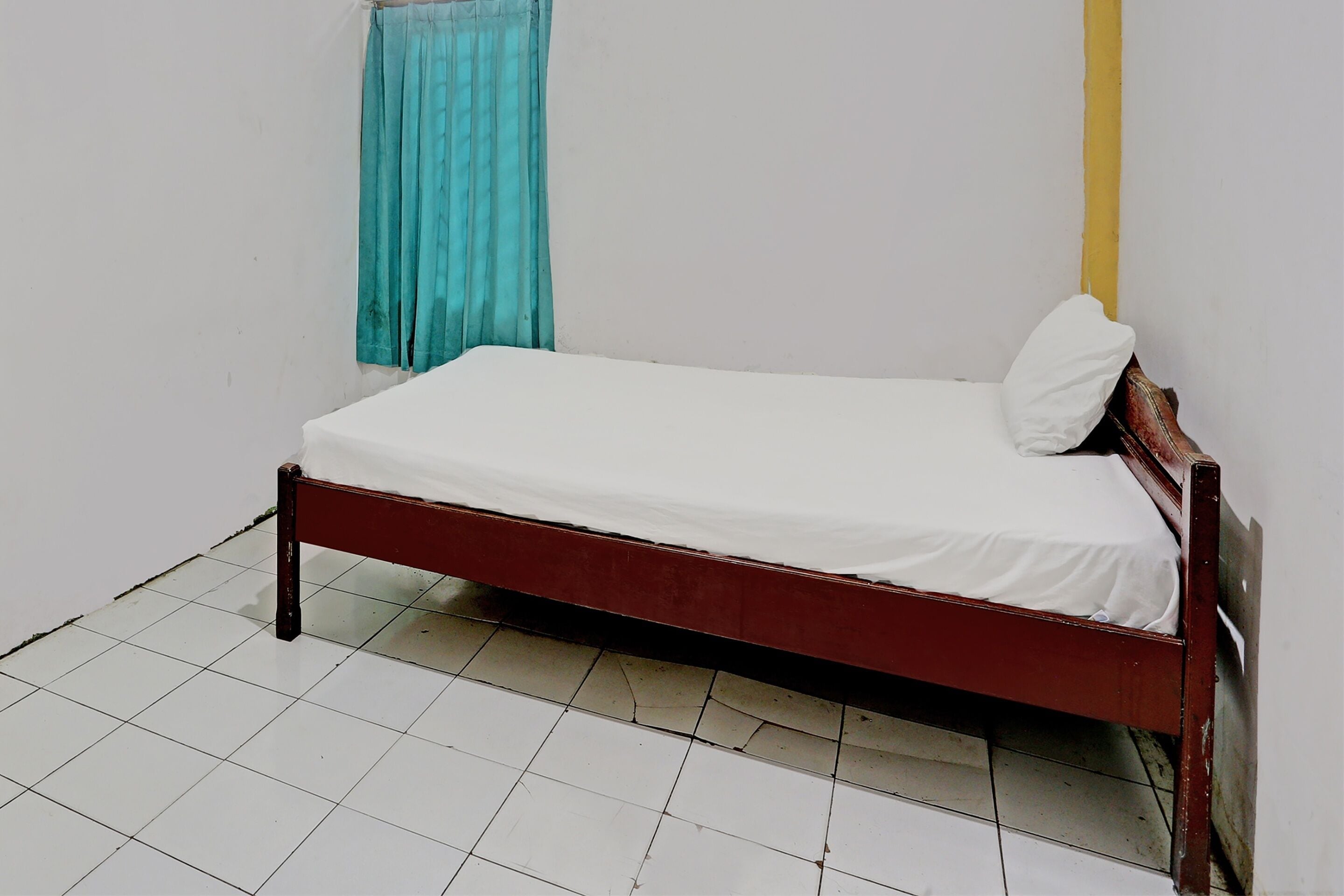 A single bed with a white mattress and a wooden frame in a room with white tiled flooring and a plain white wall.