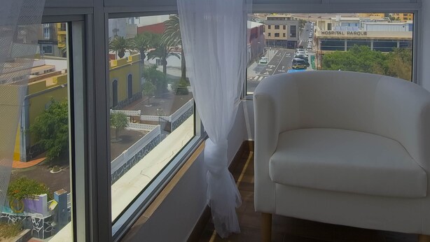 Une pièce dotée d’une grande fenêtre, d’une chaise blanche et offrant une vue sur un parc et des bâtiments d’un hôpital.
