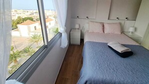 Une chambre avec une grande fenêtre, un lit, une table de chevet et une commode.