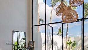 Amplia sala de estar con grandes ventanales, una silla de madera, una mesa redonda y vistas a una piscina y palmeras.