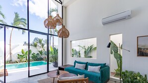 Un salón moderno con un sofá azul aguamarina, una silla de madera y amplios ventanales con vistas a una piscina y plantas tropicales.