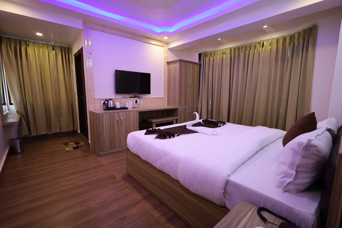 A hotel room with a large bed, a flat-screen TV mounted on the wall, wooden cabinets, and a small table with a kettle and cups.