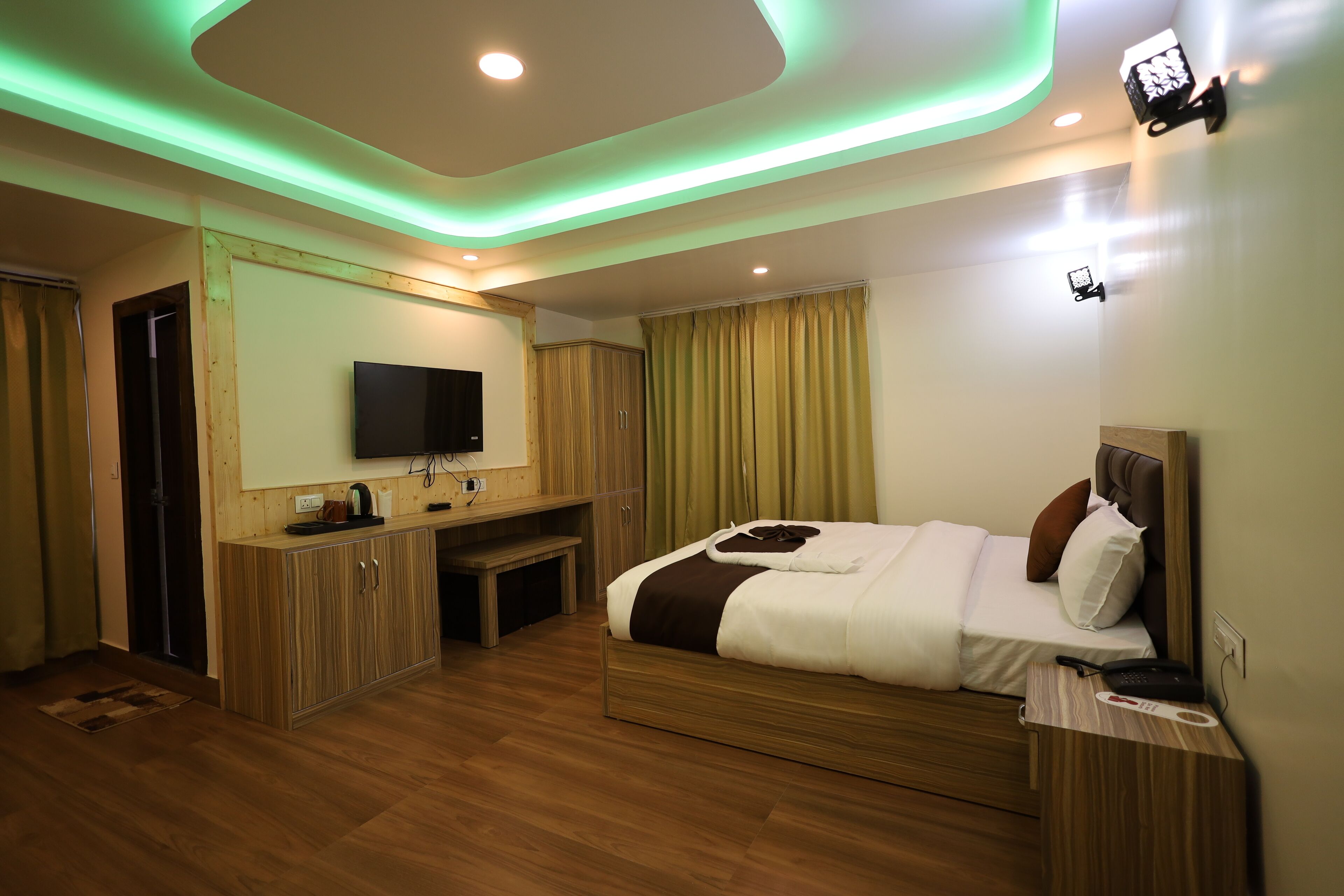 A modern hotel room with a wooden floor, a bed with white bedding, a TV mounted on the wall, and a desk with a chair.