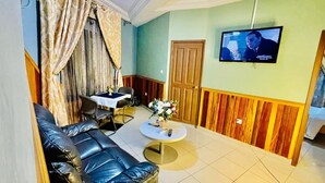 A room with a black leather sofa, a small round table with a vase of flowers, a wooden door, and a television mounted on the wall. - MARVEL HOTEL BUEA (Buea)