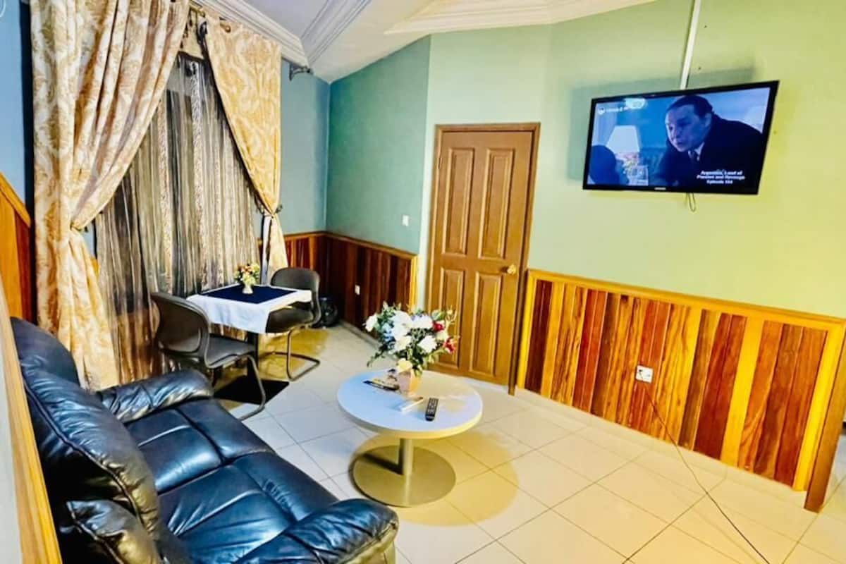 A room with a black leather sofa, a small round table with a vase of flowers, a wooden door, and a television mounted on the wall.