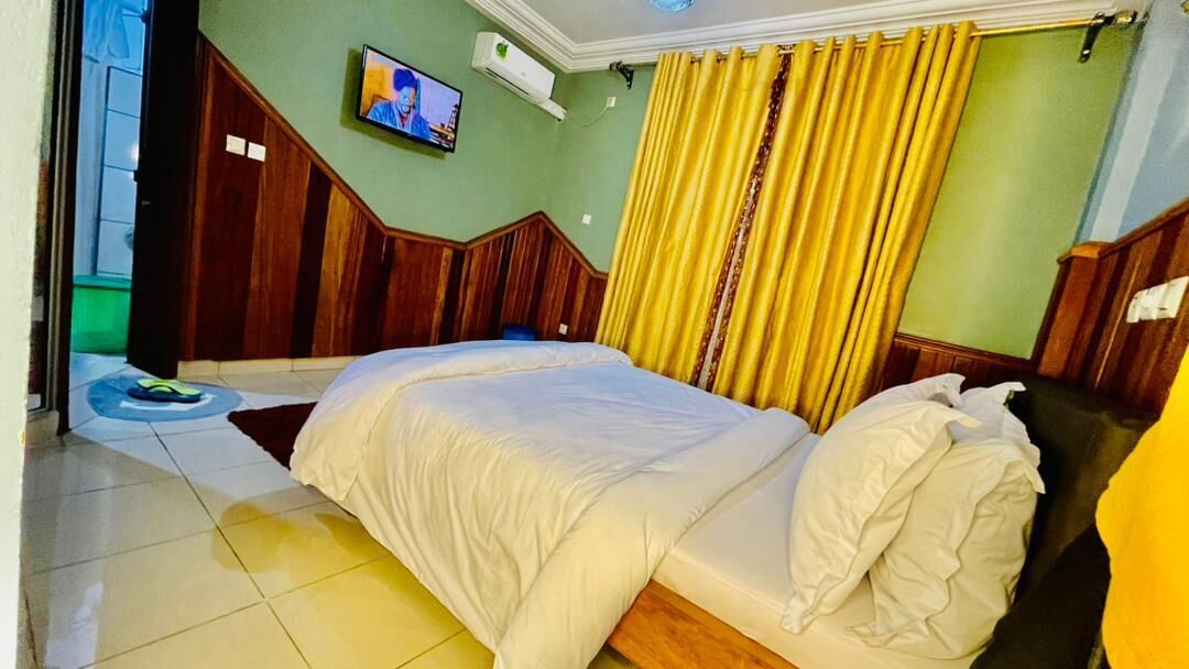 Une chambre d’hôtel avec un grand lit, une tête de lit en bois, un téléviseur fixé au mur et des rideaux jaunes.