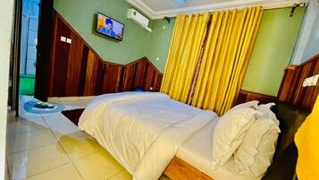 Une chambre d’hôtel avec un grand lit, une tête de lit en bois, un téléviseur fixé au mur et des rideaux jaunes.