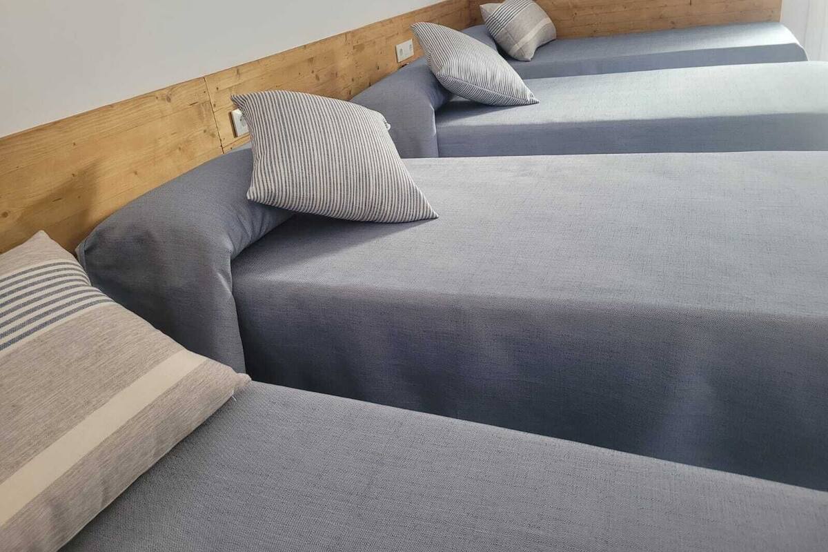 Una esquina de una habitación con tres camas, cada una con cubrecama gris y almohadas a rayas.